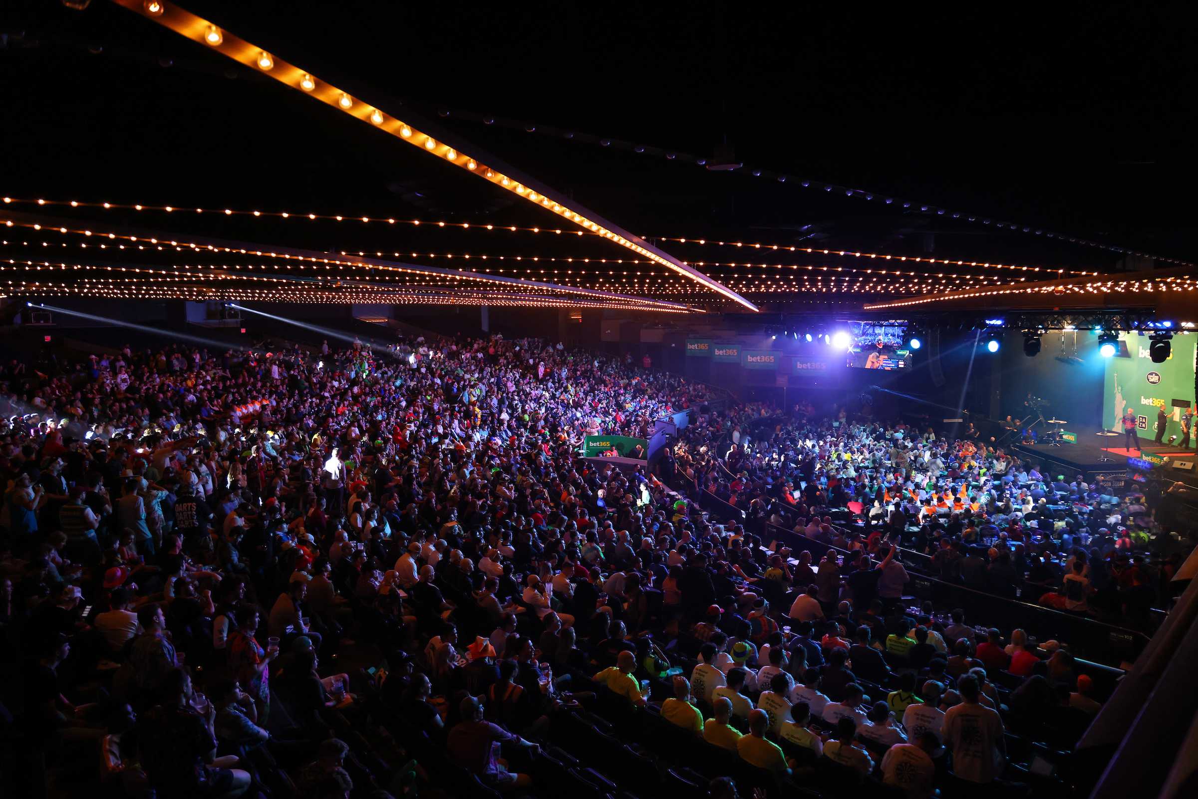 A packed arena watching a professional darts match under festoon lights